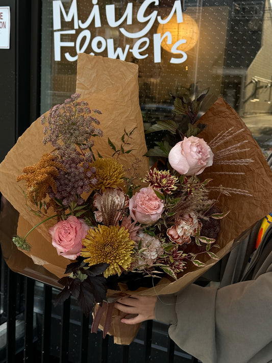 Bouquet featuring pink roses, yellow chrysanthemums, and dried flowers wrapped in kraft paper, held outside Miugi Flowers shop.
