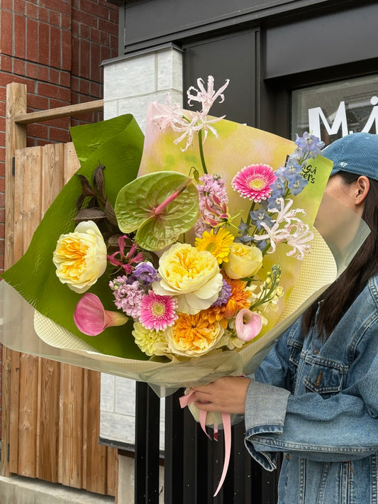 A person holding a bouquet featuring yellow garden roses, pink ranunculus, green anthurium, orange marigolds, pink gerbera daisies, purple stock flowers, blue delphinium, and pink nerine. The bouquet is wrapped in light green and beige paper with pink ribbon, and the background includes a wooden fence and brick building.
