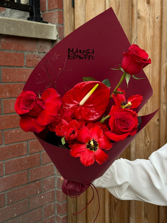A hand holding a red floral bouquet wrapped in deep red paper with the “Miugi Flowers” logo. The bouquet features red roses, red anthurium, and vibrant red tulips, creating a monochromatic red arrangement with lush greenery as accents. The background includes a brick wall and wooden panels.