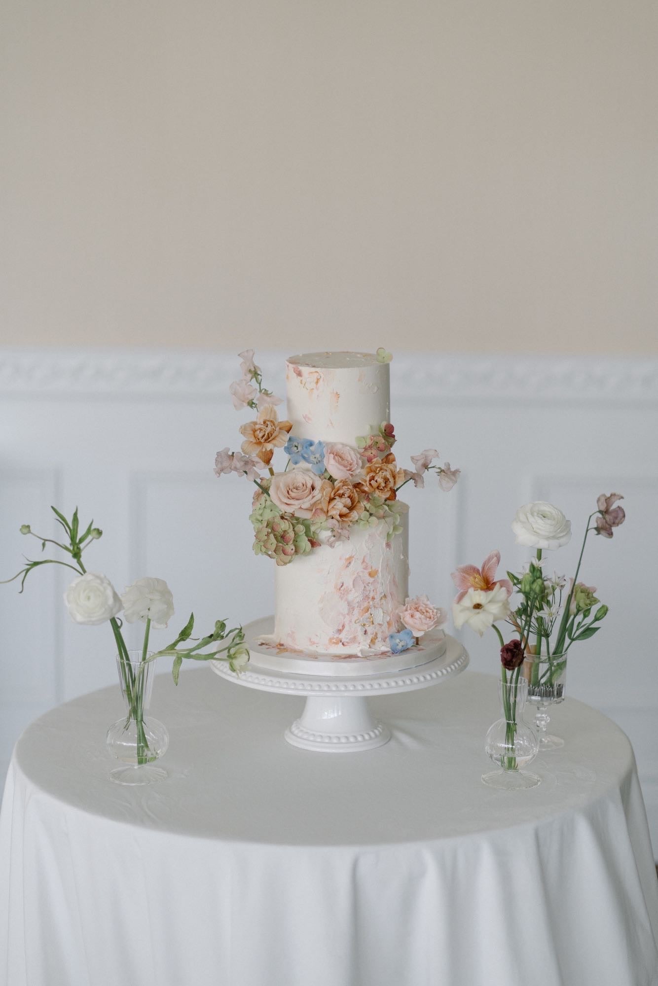 A two-tiered white wedding cake decorated with blush pink, peach, and blue flowers, placed on a white cake stand. The cake is on a round table covered with a white tablecloth. Surrounding the cake are small glass vases holding single stems of white ranunculus, lisianthus, and dark-colored flowers, set against a neutral wall with white wainscoting.