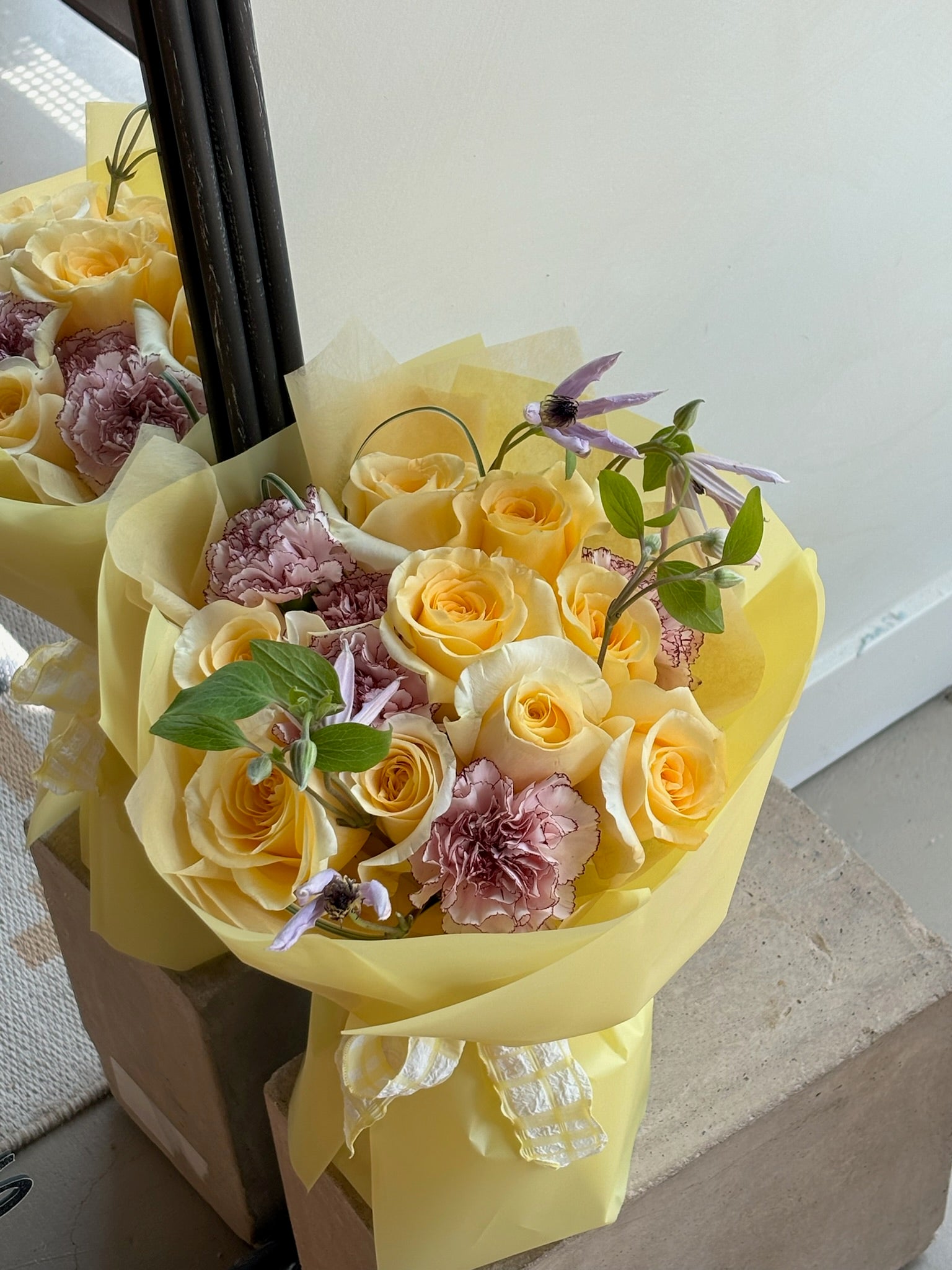 A bouquet featuring bright yellow roses, light purple carnations, and delicate purple accent flowers with greenery. The arrangement is wrapped in matching yellow paper and tied with a ribbon, placed on a concrete surface near a mirror reflecting the bouquet. The setting is indoors with natural lighting.